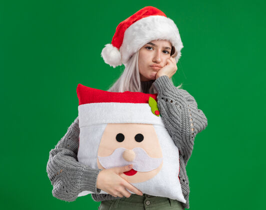 快乐年轻的金发女郎穿着冬衣 戴着圣诞帽 抱着圣诞枕头 站在绿色的背景下 看着镜头 困惑和不快圣诞快乐不高兴抱着