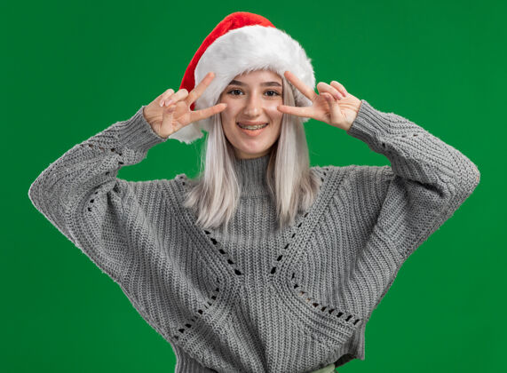圣诞年轻的金发女郎穿着冬季毛衣 戴着圣诞帽 看着摄像机 兴高采烈地微笑着 绿色背景上立着v字标志圣诞老人金发碧眼微笑