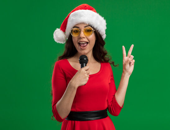 圣诞快乐的年轻漂亮女孩戴着圣诞帽戴着眼镜拿着麦克风看着相机在绿色背景上做着和平的标志和复制空间隔离快乐快乐眼镜