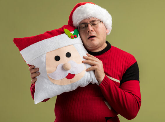 圣诞老人昏昏欲睡的成年男子戴着眼镜 戴着圣诞帽 抱着圣诞老人的枕头 抚摸着它的头 在橄榄绿的背景上与世隔绝地睡觉触摸快乐头