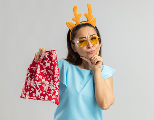 女人身穿蓝色上衣的年轻女子 戴着滑稽的鹿角边 戴着黄色眼镜 手里拿着圣诞红包 手放在下巴上思考着眼镜快乐顶