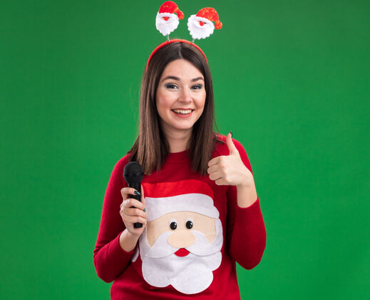 圣诞老人快乐的年轻漂亮的白人女孩穿着圣诞老人的毛衣和头带拿着麦克风显示拇指向上隔离在绿色墙壁与复制空间圣诞节拇指圣诞老人