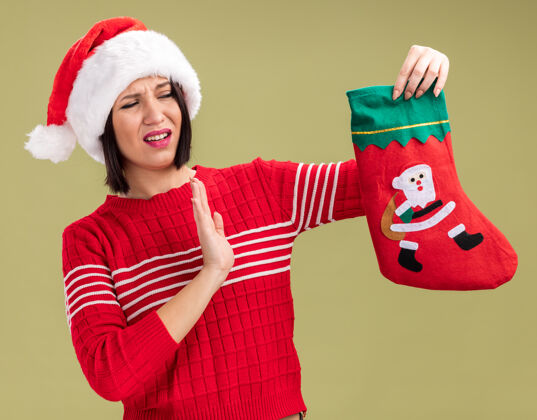 圣诞老人恼怒的年轻女孩戴着圣诞帽拿着圣诞长袜看着它在橄榄绿的墙上做着拒绝的手势拒绝穿长袜