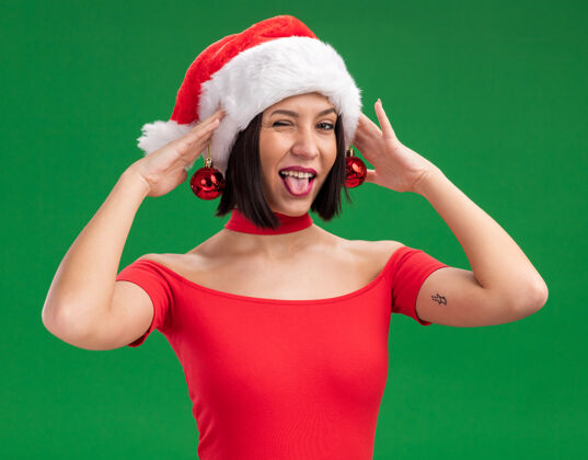 穿着可爱的小女孩戴着圣诞帽拿着圣诞饰品挂在耳朵上 舌头眨着眼睛孤立地站在绿色的墙上圣诞饰品舌头帽子