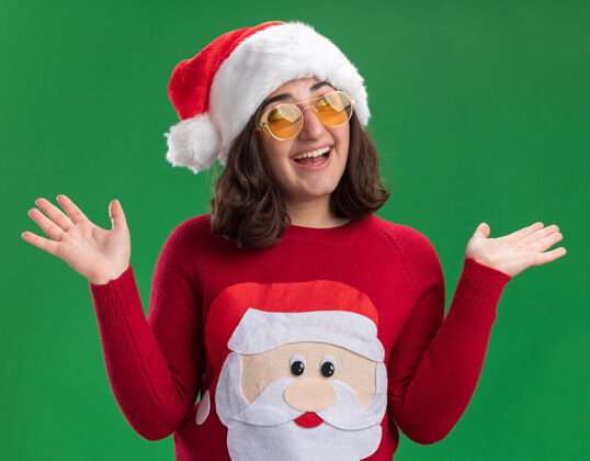 举起穿着圣诞毛衣 戴圣诞帽 戴着圣诞眼镜的年轻女孩站在绿色的墙上 开心地抬起手来看着一边帽子放在一边毛衣