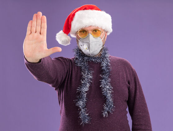 圣诞节严苛的中年男子戴着圣诞帽 戴着防护面具 脖子上戴着金属丝花环 戴着眼镜 做着停止的手势 隔离在紫色的墙上圣诞老人停止做