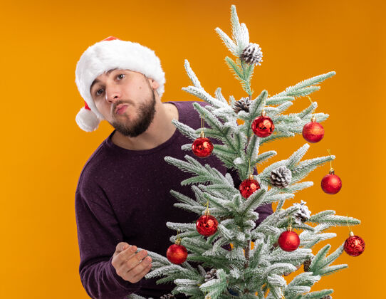 男人一个身穿紫色毛衣 戴着圣诞帽 满脸幸福的年轻人站在橙色墙上的圣诞树旁站着圣诞老人毛衣