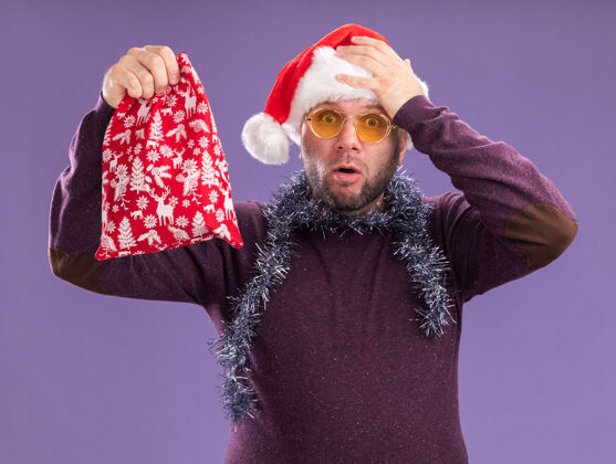 男人遗憾的是 中年男子戴着圣诞帽 脖子上戴着金箔花环 戴着眼镜 手里拿着圣诞礼品袋 手放在头上 被隔离在紫色的墙上穿着脖子圣诞节