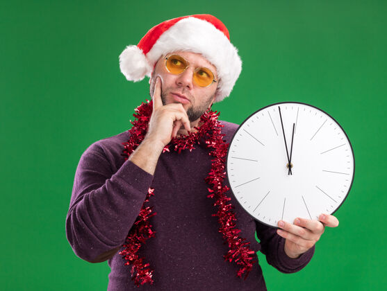 时钟体贴的中年男子戴着圣诞老人帽 脖子上戴着金箔花环 戴着眼镜 手里拿着时钟 手放在下巴上 孤零零地看着绿色的墙上周围穿着圣诞节