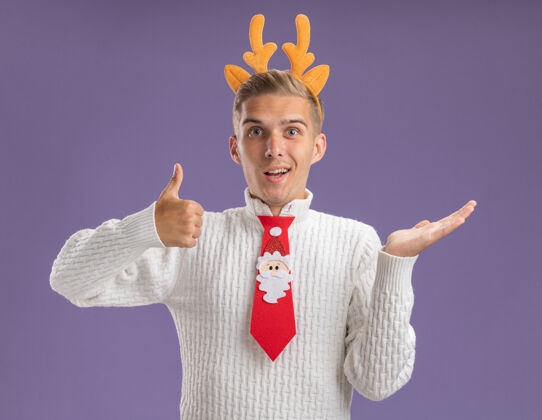 空印象深刻的年轻帅哥穿着驯鹿鹿角头带和圣诞老人领带显示空手和拇指向上孤立的紫色墙上印象深刻领带驯鹿