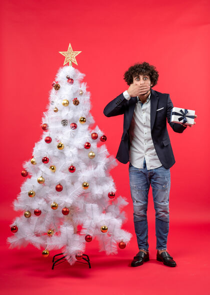 身边震惊的年轻人拿着他的礼物 闭上了嘴站在红色的右侧装饰的白色圣诞树旁对年轻人圣诞
