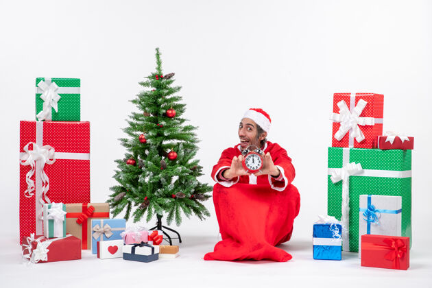 快乐年轻人坐在地上庆祝圣诞节 并在礼物和圣诞树旁展示钟表男人装饰装饰