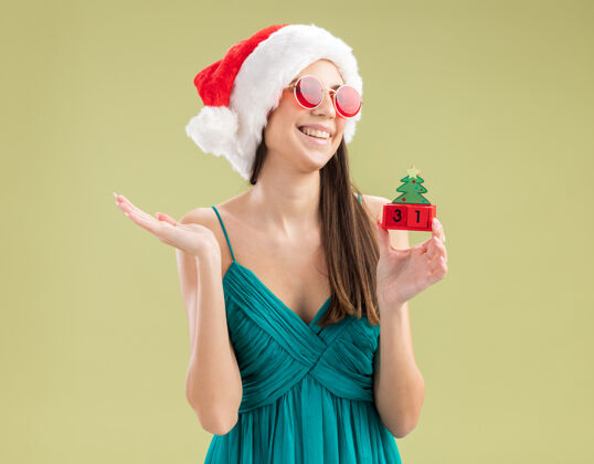 侧快乐的年轻白种女孩戴着太阳眼镜 戴着圣诞帽 拿着圣诞树的装饰品 看着身边橄榄圣诞老人圣诞树