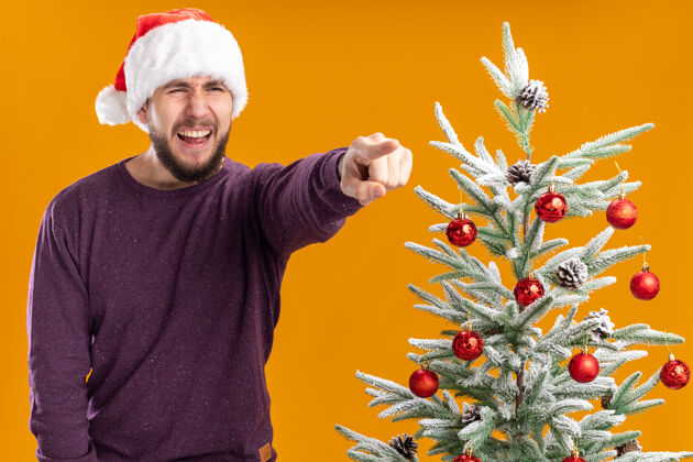帽子一个穿着紫色毛衣 戴着圣诞帽的年轻人 带着恼怒的表情 用食指指着站在橙色背景下圣诞树旁边的东西食指有些事男人