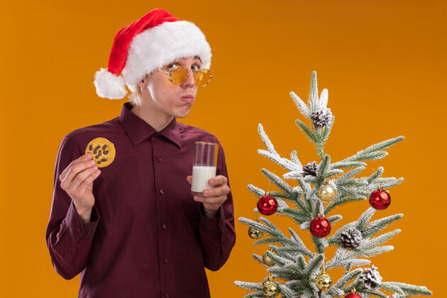 帽子困惑的年轻金发男子戴着圣诞帽 戴着眼镜 站在装饰好的圣诞树旁 手里拿着一杯牛奶和饼干 看着孤立在橙色背景上的摄像机年轻抱着靠近