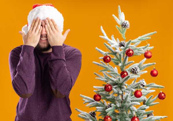 眼睛穿着紫色毛衣 戴着圣诞帽的年轻人用手遮住眼睛 旁边是橙色背景下的圣诞树圣诞树圣诞老人封面