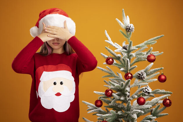眼睛穿着圣诞毛衣和圣诞帽的小女孩闭上眼睛 手放在橙色背景的圣诞树旁女孩圣诞节手