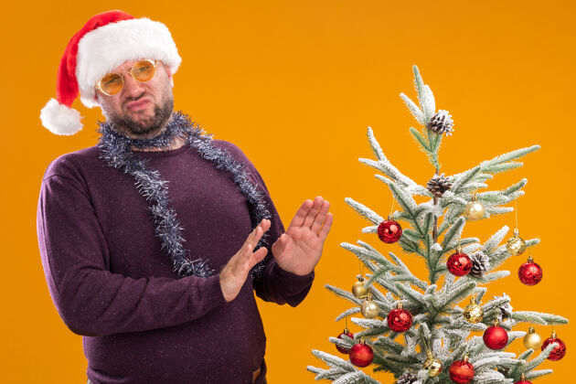 穿一个戴着圣诞帽 脖子上戴着金属丝花环 戴着眼镜的中年男子站在装饰好的圣诞树旁看着照相机站着男人脖子