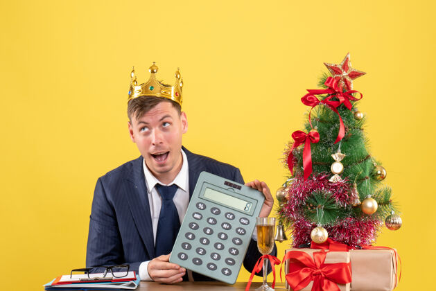 乐器商务人士手持计算器坐在圣诞树旁的桌子前 呈现黄色圣诞节持有工作