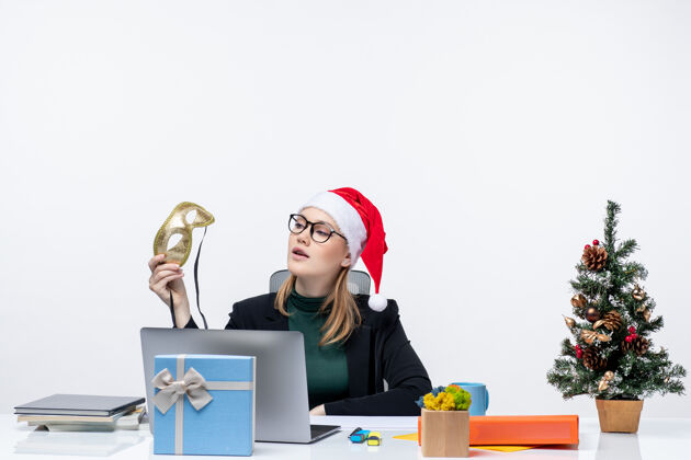 面具戴着圣诞老人帽 戴着眼镜 拿着面具的年轻女子坐在桌子旁 手里拿着圣诞树和礼物电脑桌子盒子