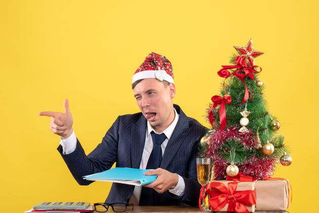 眨眼商人的正面图 眨眼的手指指着坐在圣诞树旁桌子上的人 黄色的礼物男人生意坐着