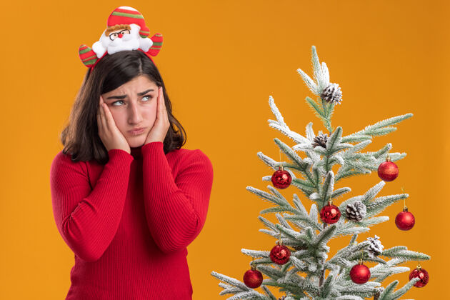 焦虑穿着圣诞毛衣的小女孩戴着滑稽的头带 困惑地看着旁边一棵橙色背景的圣诞树 非常焦虑靠边站着圣诞节
