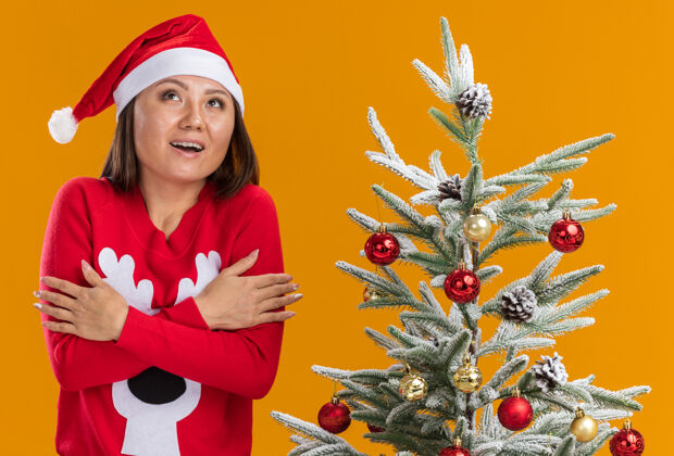 毛衣印象深刻的年轻亚洲女孩戴着圣诞帽和毛衣站在圣诞树旁交叉手孤立在橙色背景女孩在附近圣诞节