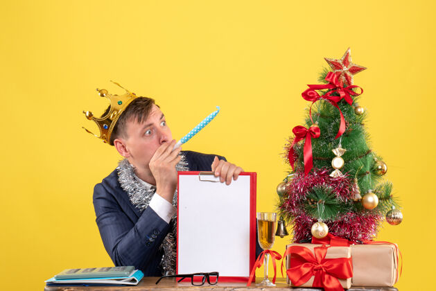 生意人前视图的商人与皇冠举行剪贴板使用noisemaker坐在桌旁圣诞树和黄色的礼物用噪音制造者生意