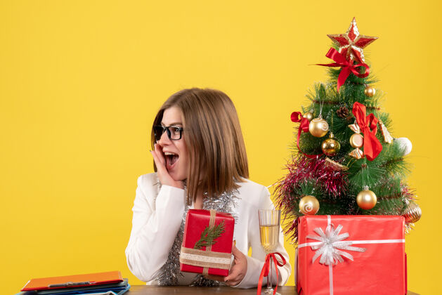 微笑正面图：女医生坐在桌子前 拿着礼物 黄色背景上的圣诞树和礼盒前面健康人