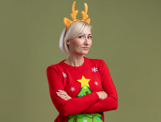 鹿角狡猾的中年金发女人戴着圣诞驯鹿鹿角头带和圣诞毛衣站在封闭的姿势看着相机在橄榄绿的背景与复制空间隔离姿势中年看