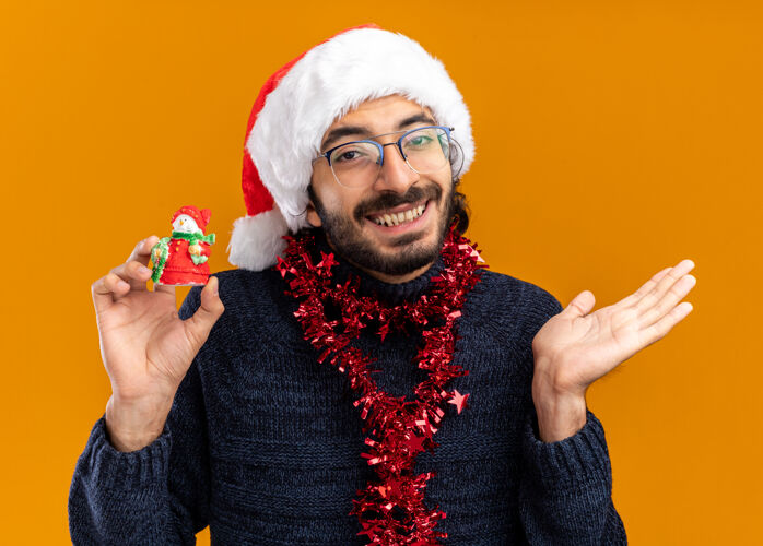 圣诞节微笑的年轻帅哥戴着圣诞帽 脖子上戴着花环 手里拿着玩具 手被隔离在橙色的背景上年轻蔓延微笑