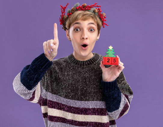 圣诞节令人印象深刻的年轻漂亮女孩戴着圣诞花环手持圣诞树玩具与日期指向隔离在紫色墙上日期印象深刻指向