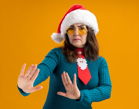 手戴着太阳眼镜 戴着圣诞帽 打着圣诞领带的未经许可的成年白人妇女双手张开 在橙色背景上没有孤立的标志 并留有复印空间未发布打开领带