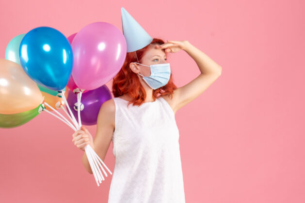 面具正面图是粉红色墙上戴着消毒面具手持彩色气球的年轻女子女性年轻女性年轻