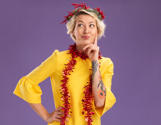 脖子体贴的金发女郎头戴圣诞花环 脖子上戴着金箔花环 手放在腰上和下巴上 在紫色背景上孤立地向上看 还有复制空间保持手金箔