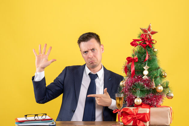 商人前视图困惑的人张开他的手坐在圣诞树附近的桌子和黄色的礼物困惑的男人秘书圣诞节