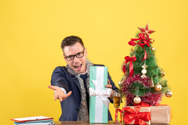 主管正面图：圣诞树旁的桌子旁 一个快乐的男人在举手 黄色的礼物快乐赠送办公室