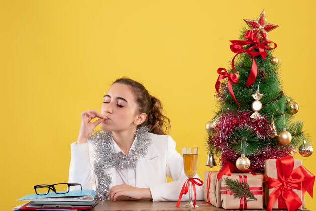 健康正面图：女医生坐在黄色背景的桌子后面 还有圣诞树和礼品盒情感快乐女医生