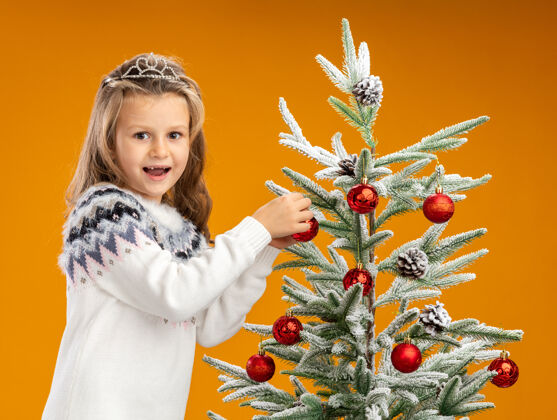 花环快乐的小女孩站在圣诞树旁边 戴着头饰 脖子上戴着花环 把树隔离在橙色的墙上戴着小脖子
