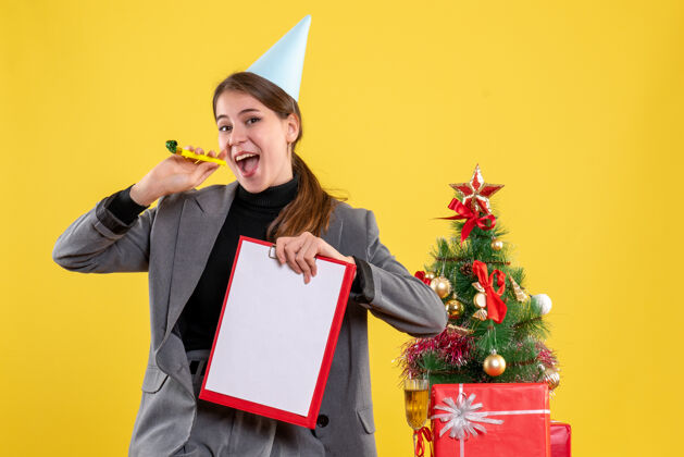 圣诞节正面图：戴着派对帽的年轻女孩拿着噪音器和文件站在圣诞树和礼物鸡尾酒旁附近知识分子年轻女孩
