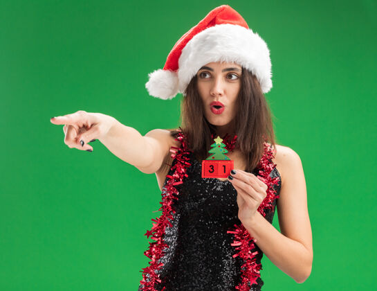 边惊讶的年轻漂亮女孩戴着圣诞帽 脖子上戴着花环 把圣诞玩具点放在隔离的绿色墙上脖子花环穿