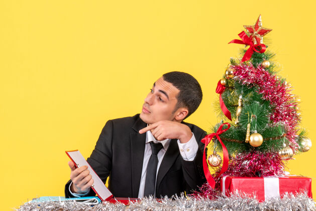 看正面图西装革履的男人坐在桌子旁看着右手拿着文件圣诞树和礼物圣诞树新郎桌子