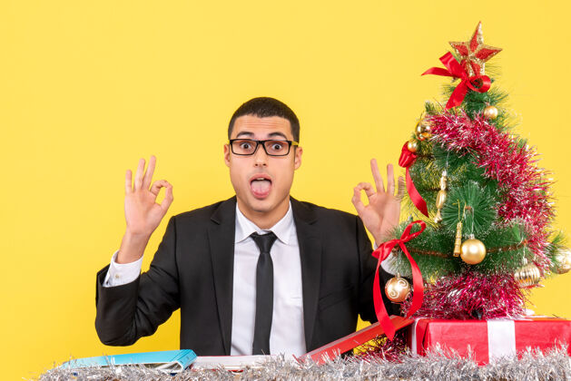 桌子正面图：张大嘴巴的年轻人坐在桌子旁制作圣诞树和礼物年轻人商人圣诞节