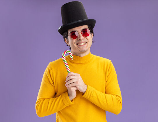 手杖穿着黄色高领毛衣戴着眼镜戴着黑色帽子拿着糖果手杖脸上带着微笑站在紫色的墙上的快乐的年轻人年轻人微笑糖果