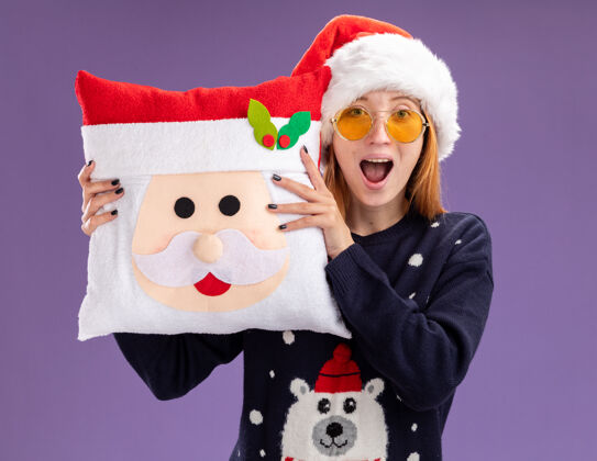 枕头惊讶的年轻漂亮女孩穿着圣诞毛衣和帽子戴着眼镜拿着圣诞枕头隔离在紫色的背景上惊喜眼镜帽子