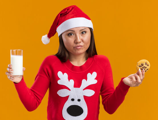 圣诞节未出嫁的年轻亚洲女孩戴着圣诞帽 穿着毛衣 手里拿着一杯牛奶 橘黄色的墙上隔着饼干未出租穿着毛衣