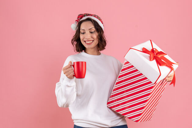 情感年轻女子手持圣诞礼物和一杯茶在粉红色墙上的前视图人茶粉色