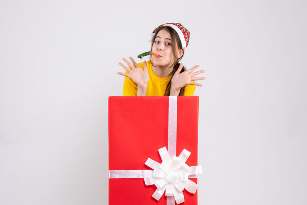 购物者戴着圣诞帽的快乐女孩站在白色大圣诞礼物后面用隔音器站立快乐大