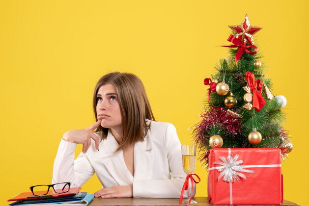 女性正面图：女医生坐在桌子前 在黄色背景上思考着圣诞树和礼品盒坐着人医院
