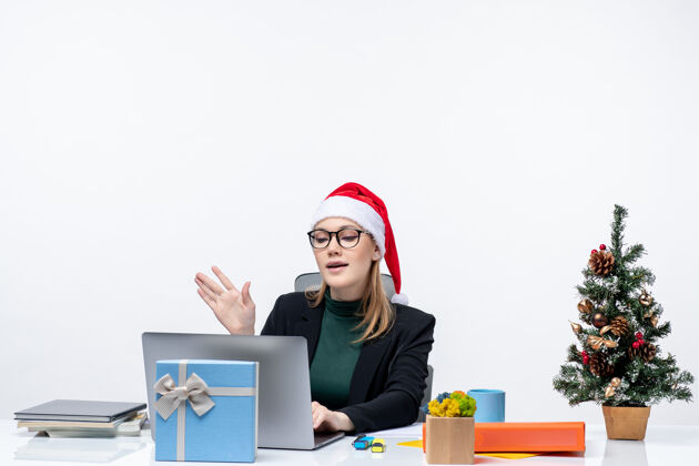 自由职业者戴着圣诞老人帽的女商人坐在桌子旁 桌上放着圣诞树和礼物商务女人圣诞老人礼物
